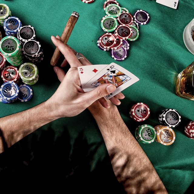 Ett par händer som håller i spelkort och en cigarr över ett bord med spelmarker och ett glas sprit.